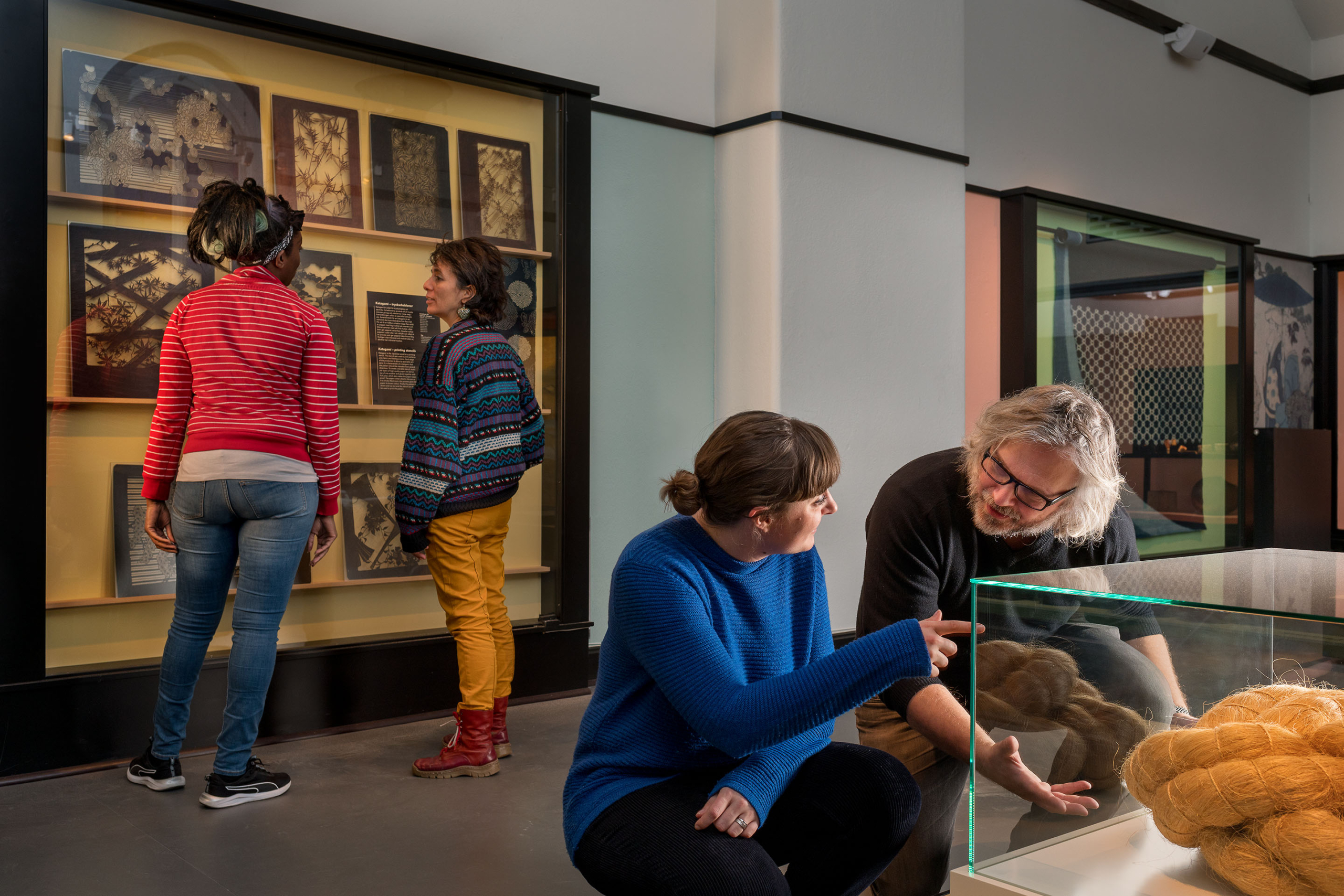 I förgrunden en kvinna i blå tröja och en man med grått hår sitter på huk framför en låg monter, i bakgrunden två mörkhåriga kvinnor vid en vägg med museiföremål.